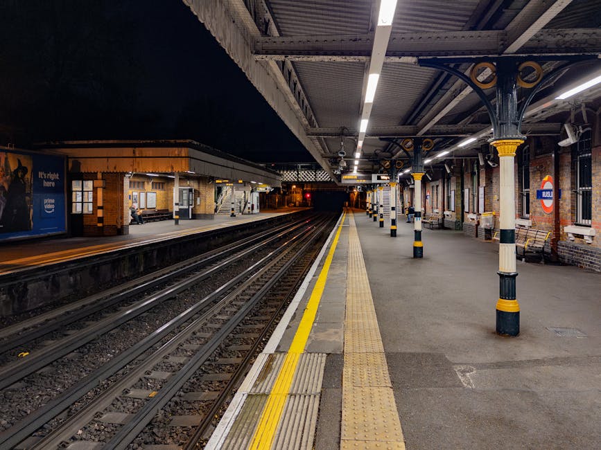 A nighttime view of an empty London Underground railway station platform featuring brick walls and metal columns painted with yellow and black stripes. The platform is illuminated by overhead lighting, with a polished concrete surface and tactile paving strips near the edge for safety. The railway tracks run parallel along the left side towards a dark tunnel in the distance. On the right, a series of benches and signage, including the iconic red and blue London Underground roundel, are visible. There are no people present on the platform. The environment appears clean and well-maintained, consistent with a typical urban transit station. This scene exemplifies a public transportation setting where independent or private waste disposal, like rubbish removal services, may be relevant for maintaining cleanliness during station management or upgrades, reflecting the ongoing need for efficient waste handling in busy urban infrastructure such as this. Rubbish Clearance Ruislip might provide services suitable for waste collection in similar commercial or public environments.
