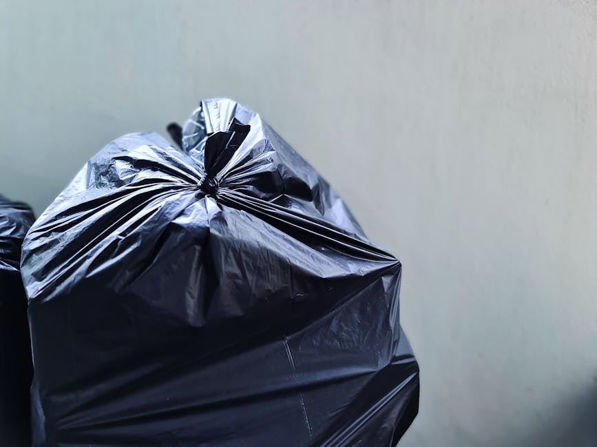 Close-up of a large black plastic rubbish bag with a shiny, crinkled texture, tightly knotted at the top, positioned in the foreground against a plain, light-colored wall. The bag appears to be full, with irregular folds and creases visible on its surface. Additional black rubbish bags are partially visible behind it, stacked or placed nearby on an indoor or outdoor surface, likely pavement or floor. The environment lacks distinguishable features but suggests a setting related to waste collection or private rubbish clearance, possibly outside a residential or commercial property. This scene aligns with services such as independent waste removal or on-site rubbish clearance, typical of companies like Rubbish Clearance Ruislip, catering to the disposal of household or commercial waste materials in a professional manner. Natural lighting highlights the reflective surface of the bag, emphasizing its fullness and material quality, with no visible damage or tears present.