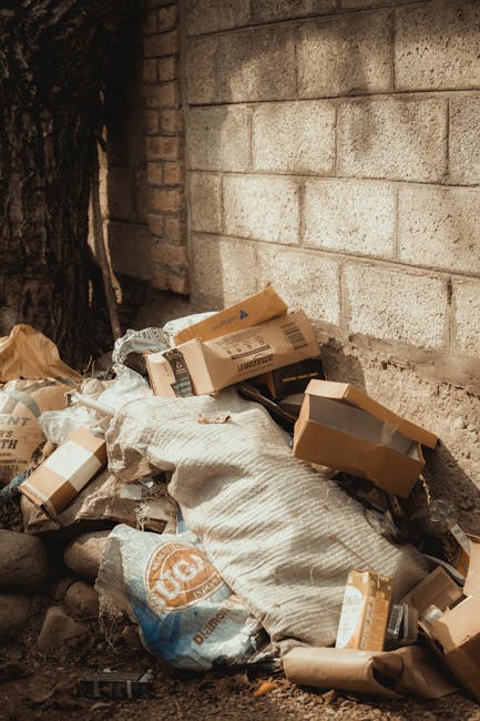 A pile of mixed household rubbish and packaging waste, including several flattened cardboard boxes, a large white sack, and plastic bags, is situated on the ground against a weathered brick wall adjacent to a tree trunk. The cardboard boxes are light brown with visible tape and printed labels, some partially crushed or torn. The white sack appears to be made of woven material, possibly containing waste, and is partially covered by the flattened boxes. The surrounding environment features an outdoor setting with a brick and concrete wall backdrop, indicating the waste is located outside a building or alongside a storage area. The ground comprises dirt and small stones, with natural sunlight illuminating the scene, highlighting the textures of the cardboard, sack, and wall. This scene illustrates a typical example of private waste or rubbish collection, where rubbish is temporarily stored before removal by a professional service like Rubbish Clearance Ruislip, emphasizing the importance of tidy and responsible disposal methods for commercial or residential waste management.