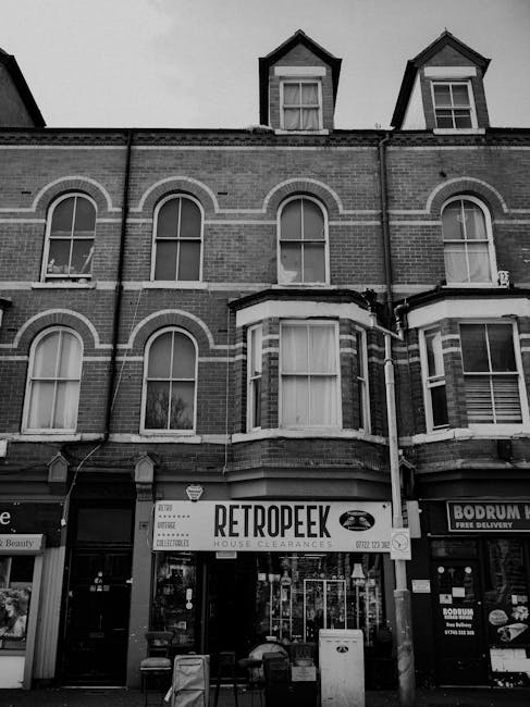 A three-story brick building with arched windows and a bay window on the ground floor, which features a sign for 'RETROPEEK' house clearance. The facade shows traditional brickwork with decorative white accents around the windows. The upper floors have sash windows, and the roof includes two small dormer windows with triangular gables. In front of the building, there is a narrow pavement area with a few small objects, possibly delivery crates or waste bins, indicating a commercial premises. To the right, part of another storefront is visible, suggesting this is situated on a busy street or shopping frontage. The scene is in black and white, emphasizing the building’s textures and architectural details, and highlighting the presence of signage that implies private or independent waste clearance services, consistent with rubbish removal themes relevant to the local area.