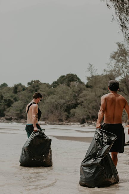 Two men are standing ankle-deep in shallow water on a beach or riverbank, each holding large black rubbish bags filled with waste. The man on the left appears to be younger, wearing dark swim trunks with a strap or backpack over his shoulder, and is looking down at the garbage bag in his hand. The man on the right is shirtless, dressed in dark shorts, and is holding a similarly filled black trash bag in his right hand while facing away from the camera. The background features a natural environment with sandy shores, a mixture of trees and shrubs, and slightly overcast skies. The scene suggests an outdoor clean-up activity, possibly organized by a waste management service such as Rubbish Clearance Ruislip, to remove rubbish from a natural or recreational area, emphasizing the importance of private disposal and local waste clearance efforts.