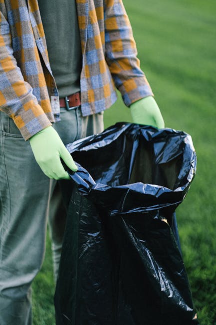 A person performing rubbish removal outdoors, standing on a grassy area. The individual is wearing a grey t-shirt, a yellow and grey checked flannel shirt, grey trousers, and bright green gloves. They are holding open a large black rubbish bag made of thick plastic, which is partially folded at the top. The background features an expanse of well-maintained green grass, suggesting a park or garden setting. The scene highlights environmental awareness and proper waste disposal, aligning with private rubbish collection services provided by companies like Rubbish Clearance Ruislip, which facilitate on-site clearance and alternative waste handling outside local authority collections.